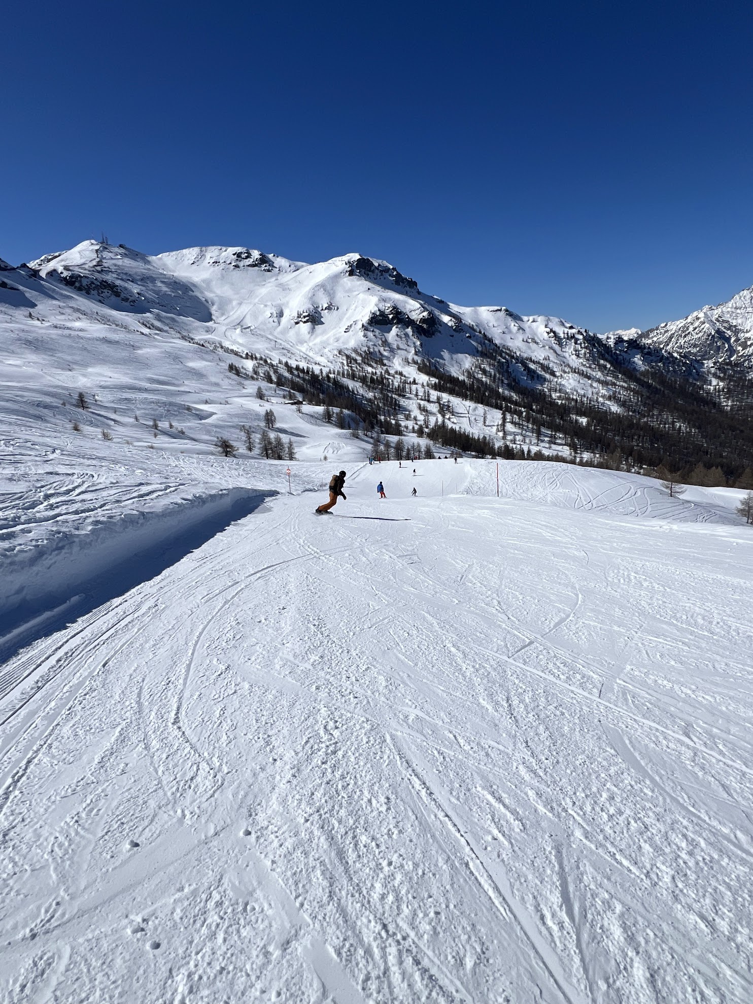 Wide view of a snowy ski slope with a snowboarder and skiers against a background of snow-covered mountains and a clear blue sky.