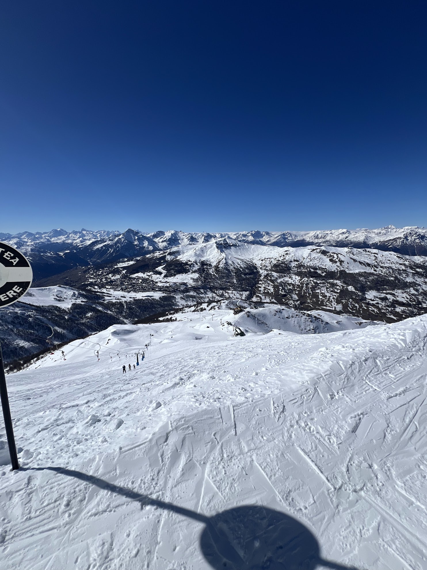 High-angle view from a snowy mountain summit looking down at a ski lift and vast snow-covered mountain ranges under a clear blue sky.