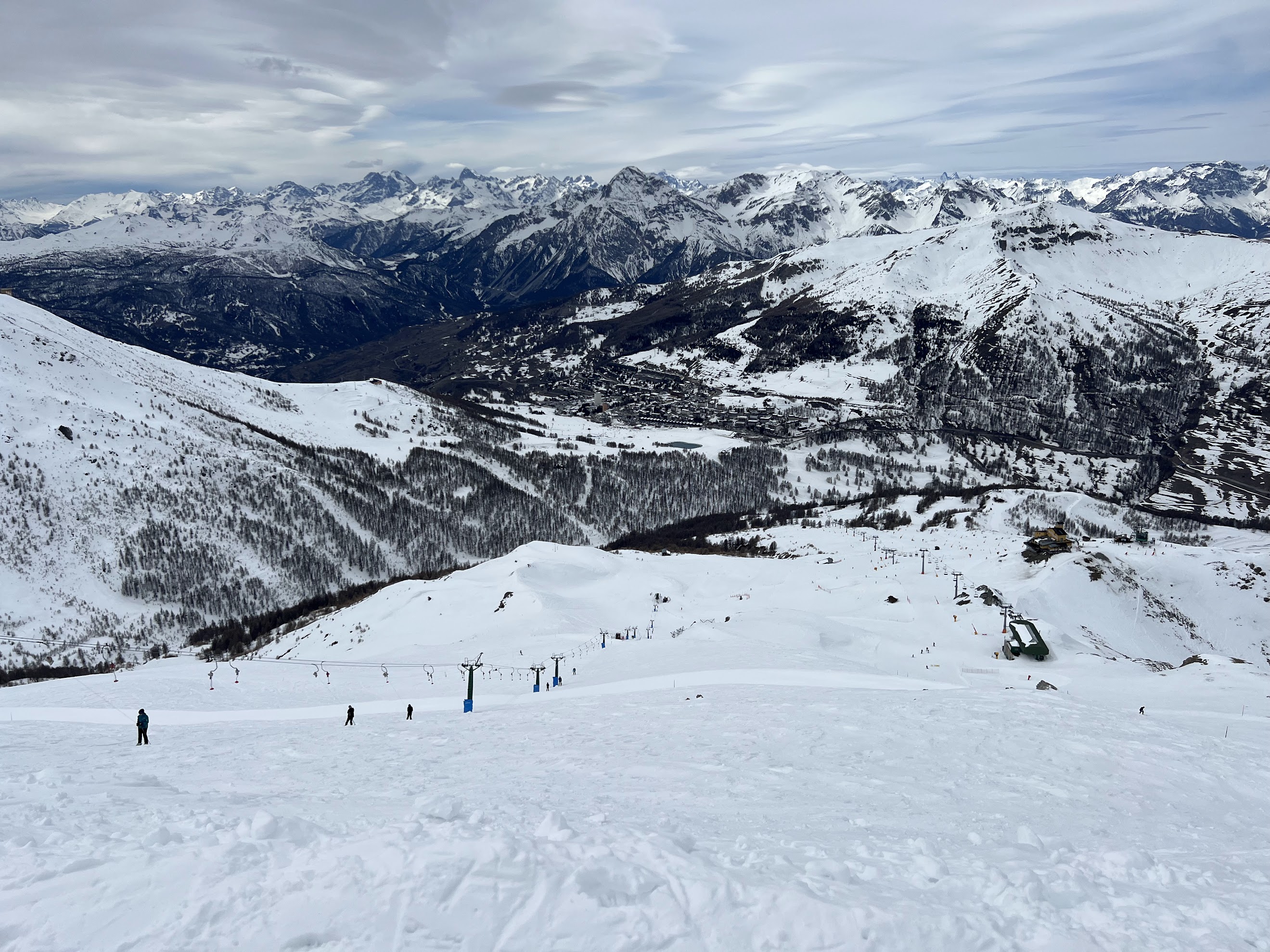 Panoramic view looking down a ski slope with ski lifts, revealing a mountain valley town surrounded by vast snow-covered alpine ranges.