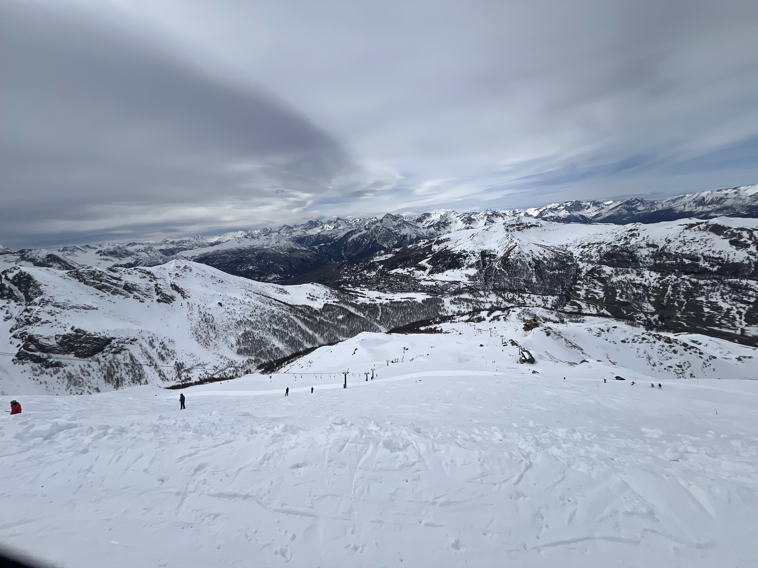 Ultra-wide panoramic view from a ski slope overlooking distant snow-covered mountains under a dramatic, cloudy sky.