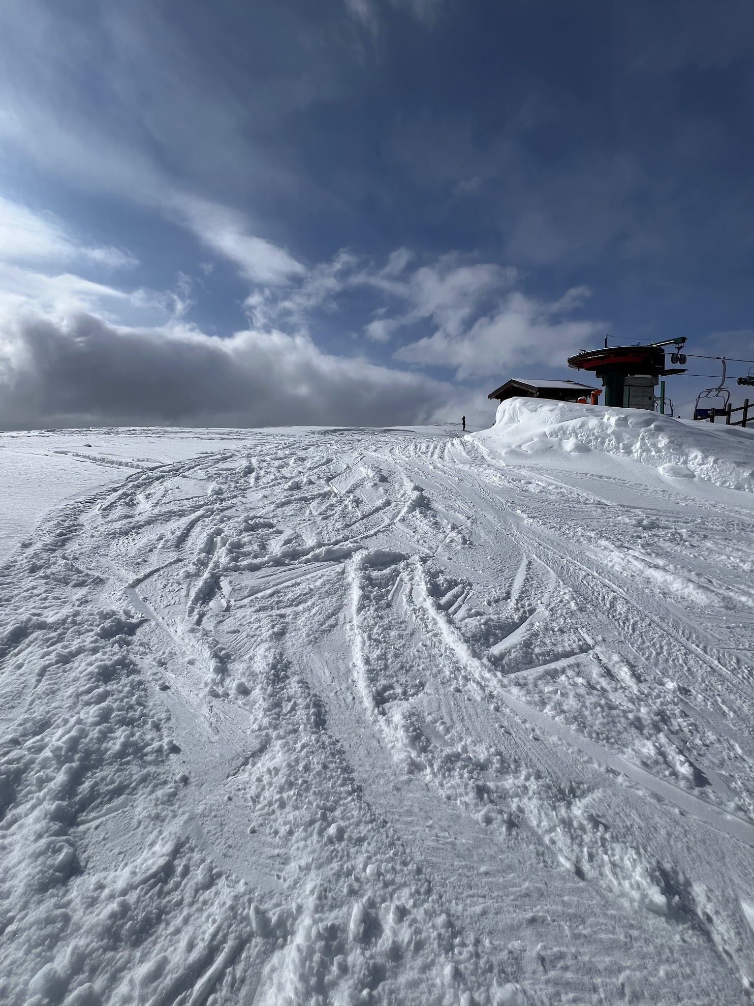Low-angle view of a snow-covered mountain peak with ski tracks, featuring a ski lift station under a cloudy blue sky.