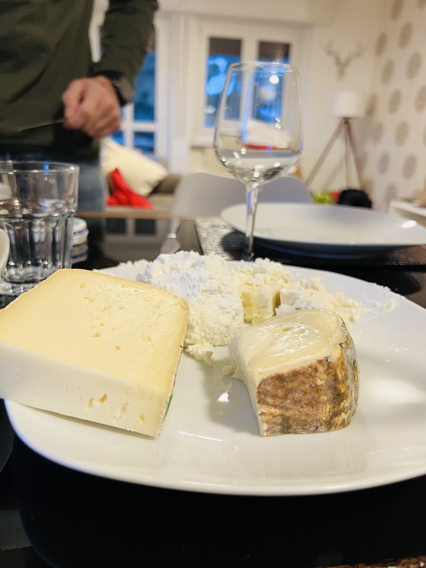 Close-up of a cheese platter on a dining table with a wine glass, plates, and a person blurred in the background.