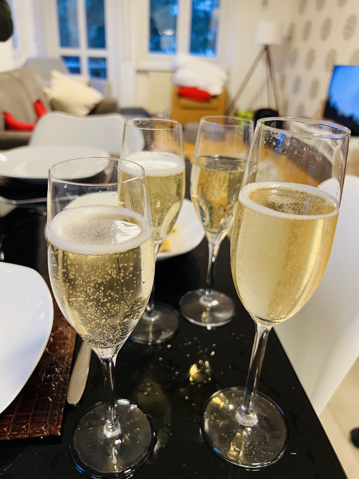Four glasses filled with sparkling wine on a dark dining table with a blurred living room background.