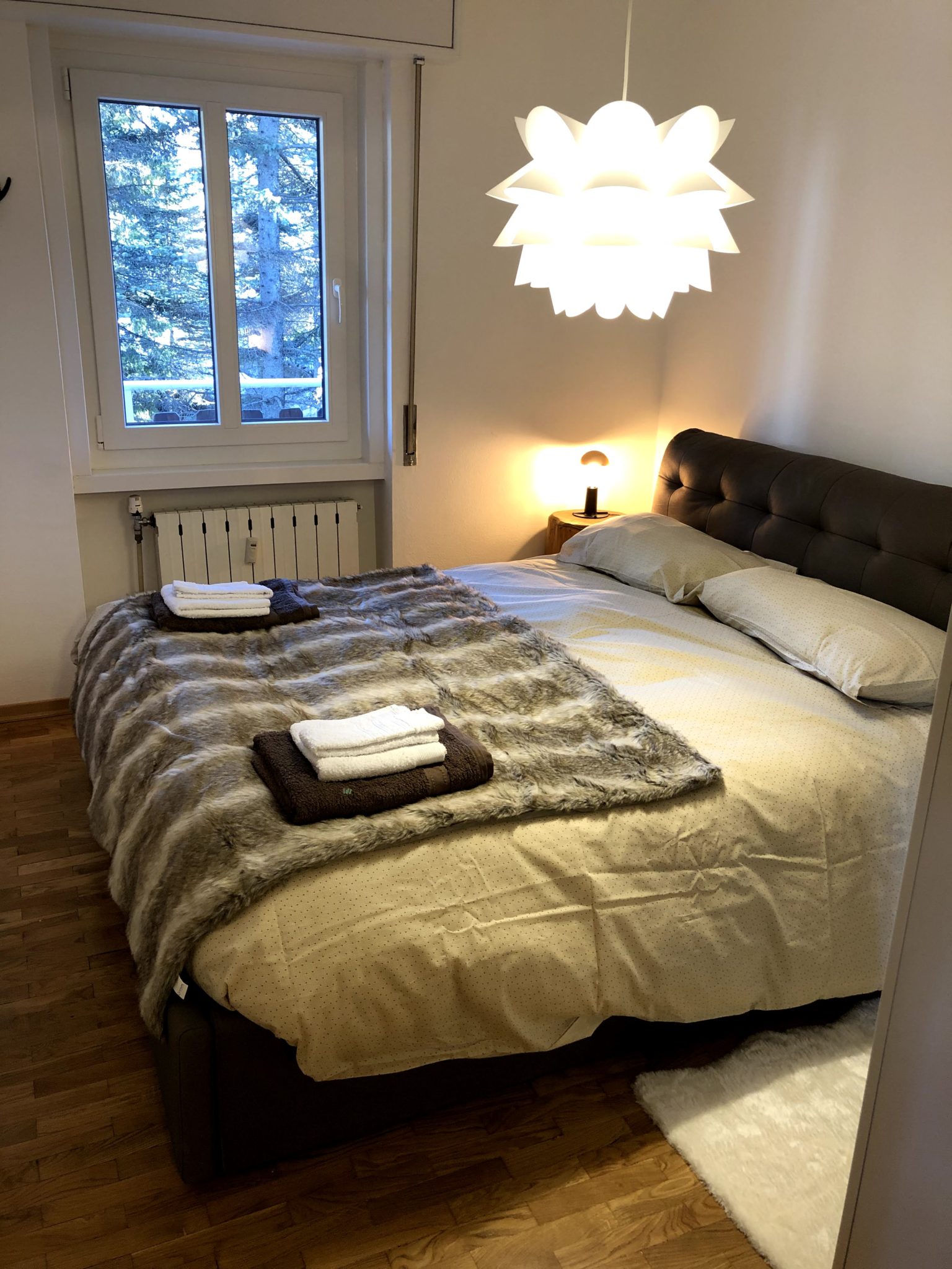 Cozy bedroom featuring a large double bed with a faux-fur throw blanket, a modern hanging light, and a window.