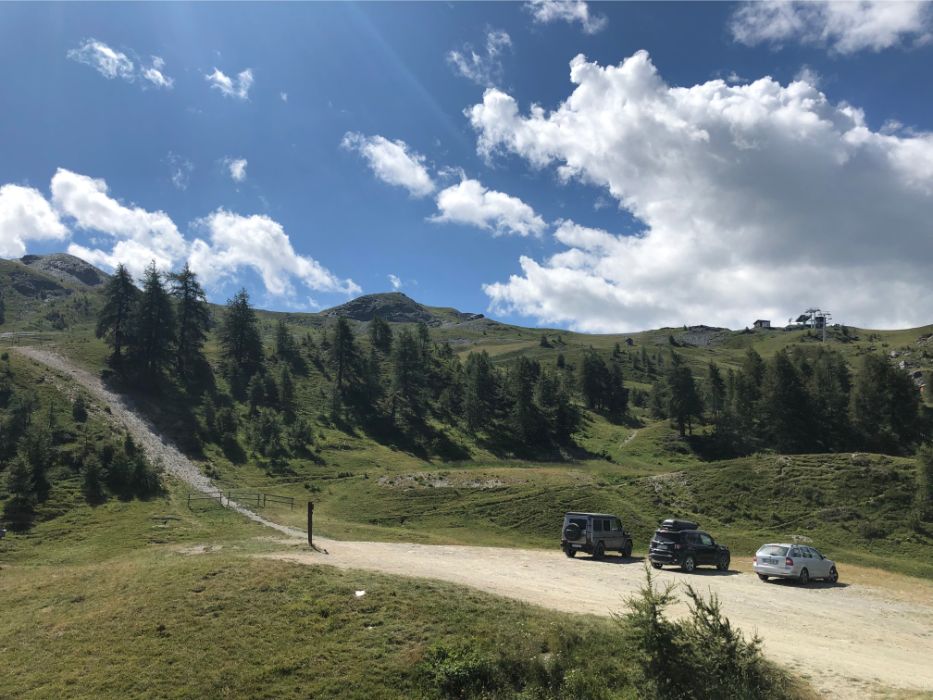 A mountain dirt parking area with three cars parked on a sunny day, surrounded by grassy green slopes and pine trees.