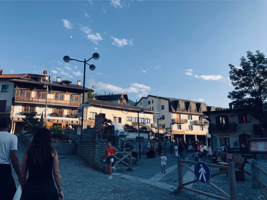 A bustling town square in an alpine resort village with traditional buildings, people walking, and a clear blue sky.
