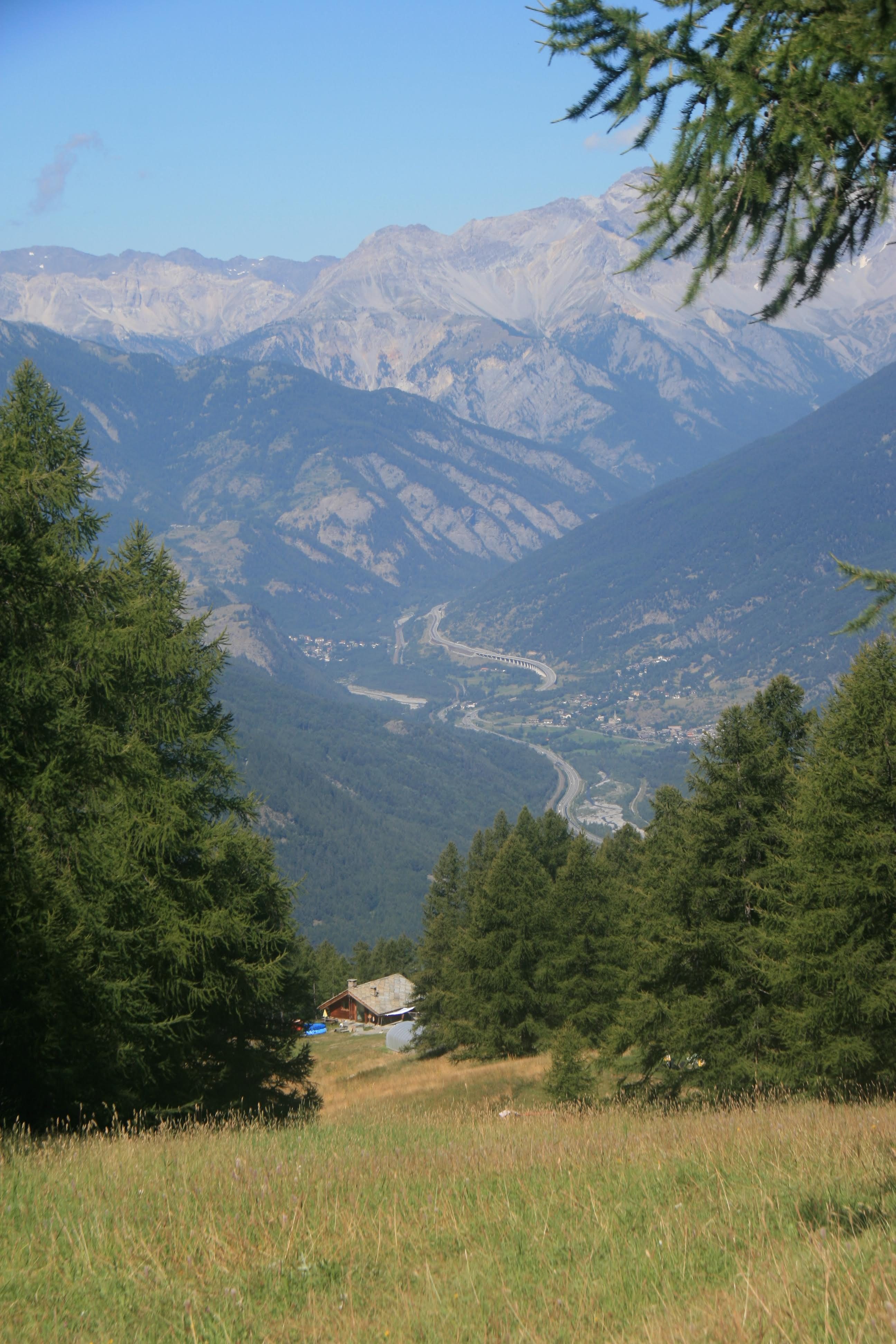 Panoramic view from a high ski resort overlooking green hills, a dirt path, a ski lift tower, and distant mountains under a cloudy sky.