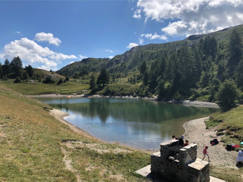 A peaceful alpine lake reflecting the surrounding green mountains and blue sky, with a stone barbecue structure in the foreground.