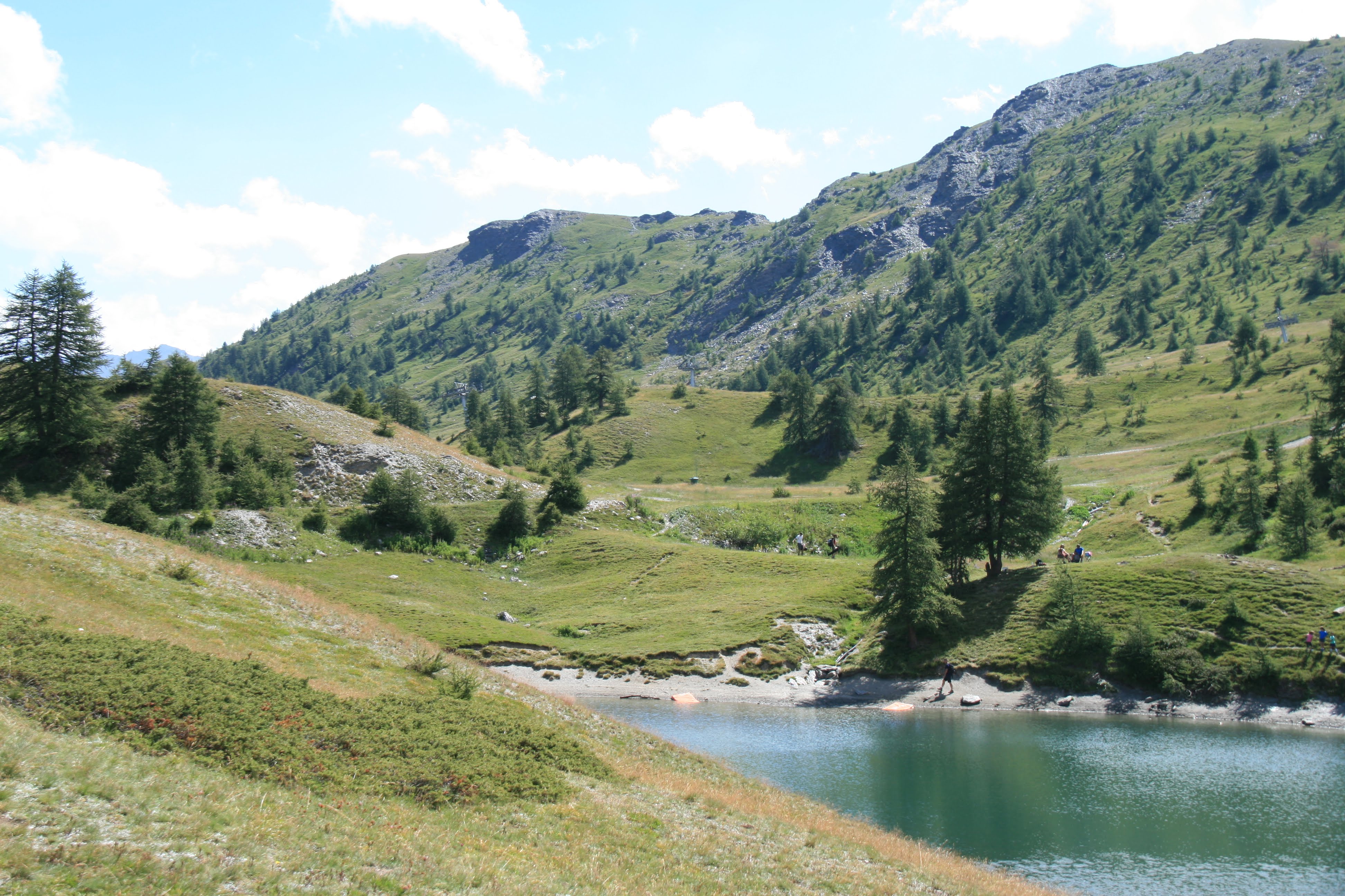 A tranquil alpine lake surrounded by rolling green hills, scattered pine trees, and a mountain peak under a partly cloudy sky.
