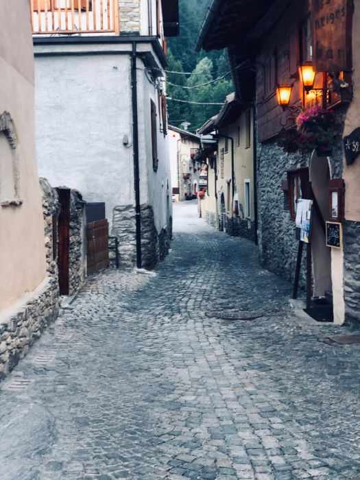A narrow cobblestone alley winding through a rustic alpine village, bordered by traditional stone and plaster houses.