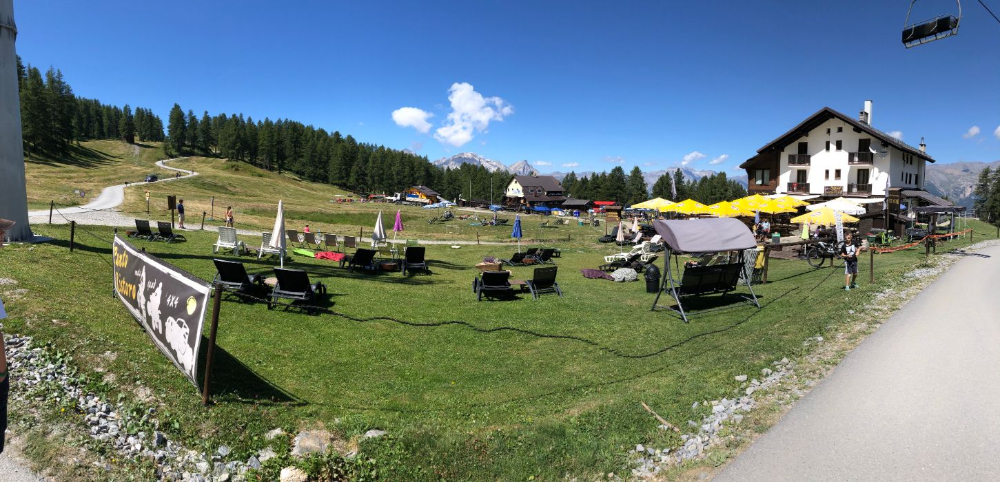 Wide panoramic view of a grassy alpine meadow with a hotel building, sun loungers, and umbrellas under a bright blue sky.