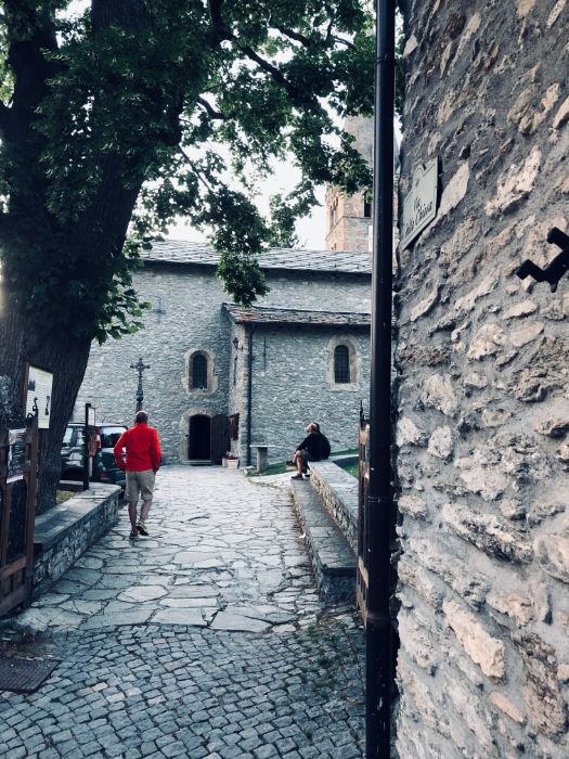 A stone path leading toward an old stone church in an alpine village, with a person in a red jacket walking in the foreground.
