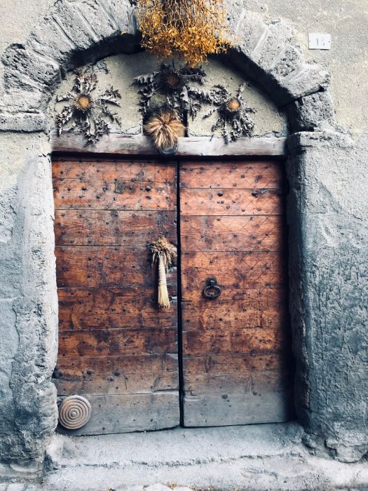 An ancient arched stone doorway with a heavy, detailed wooden door adorned with dried flowers above the frame.