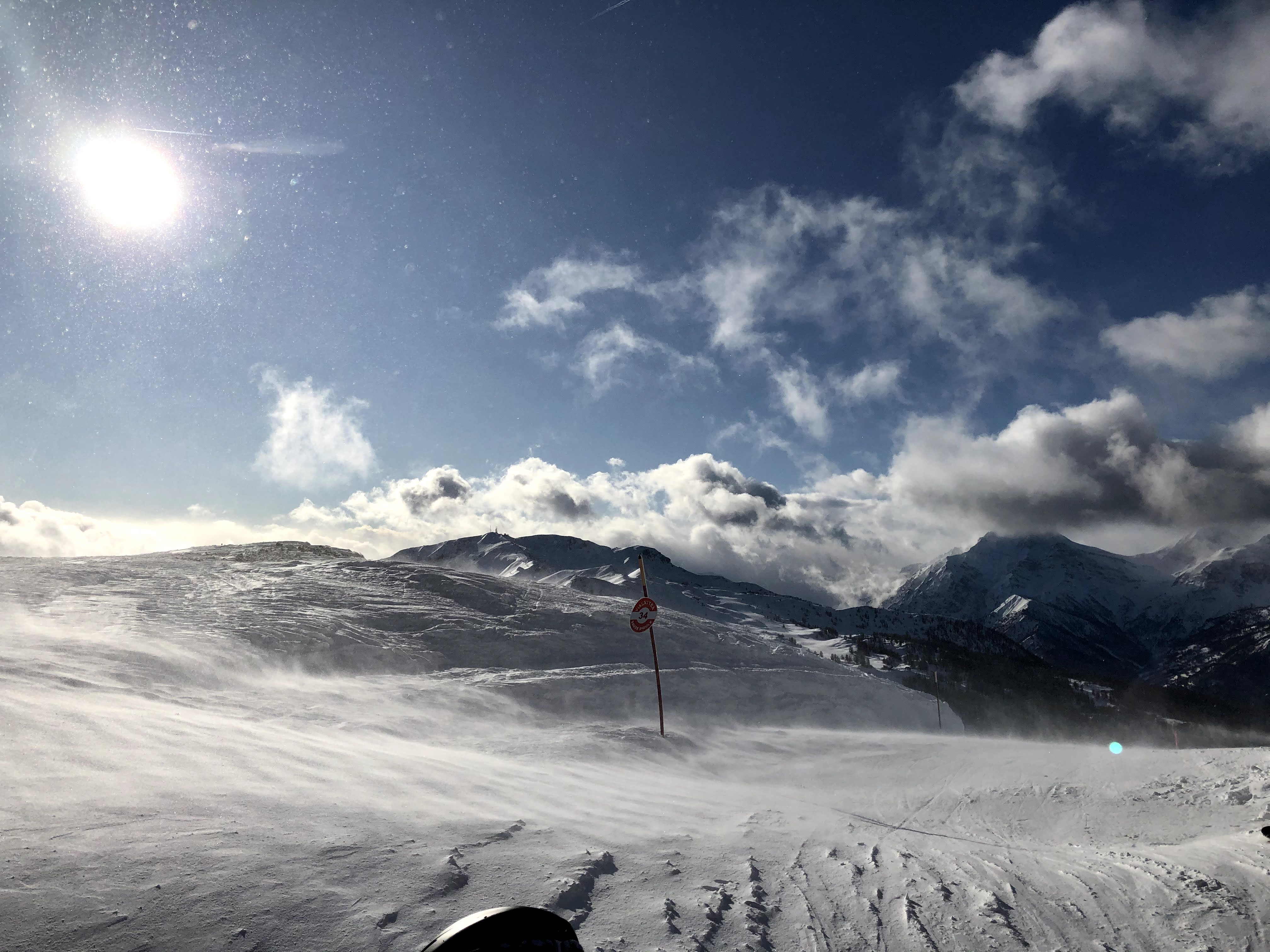 Windswept snowy mountain peak with snow blowing across the ground under a bright sun and partly cloudy sky.
