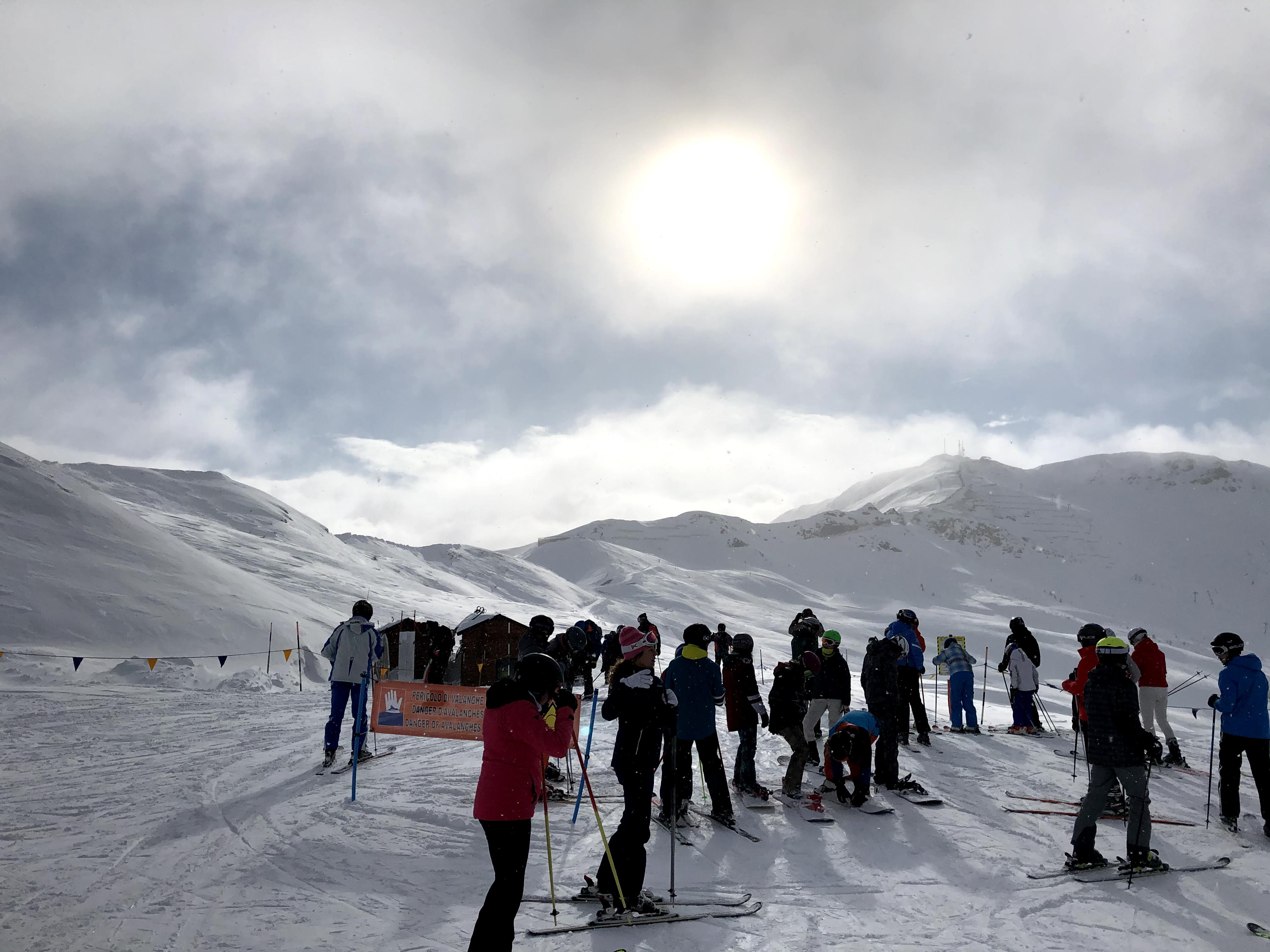 A group of skiers preparing on a snowy slope under a hazy sun, with mountains rising in the background.