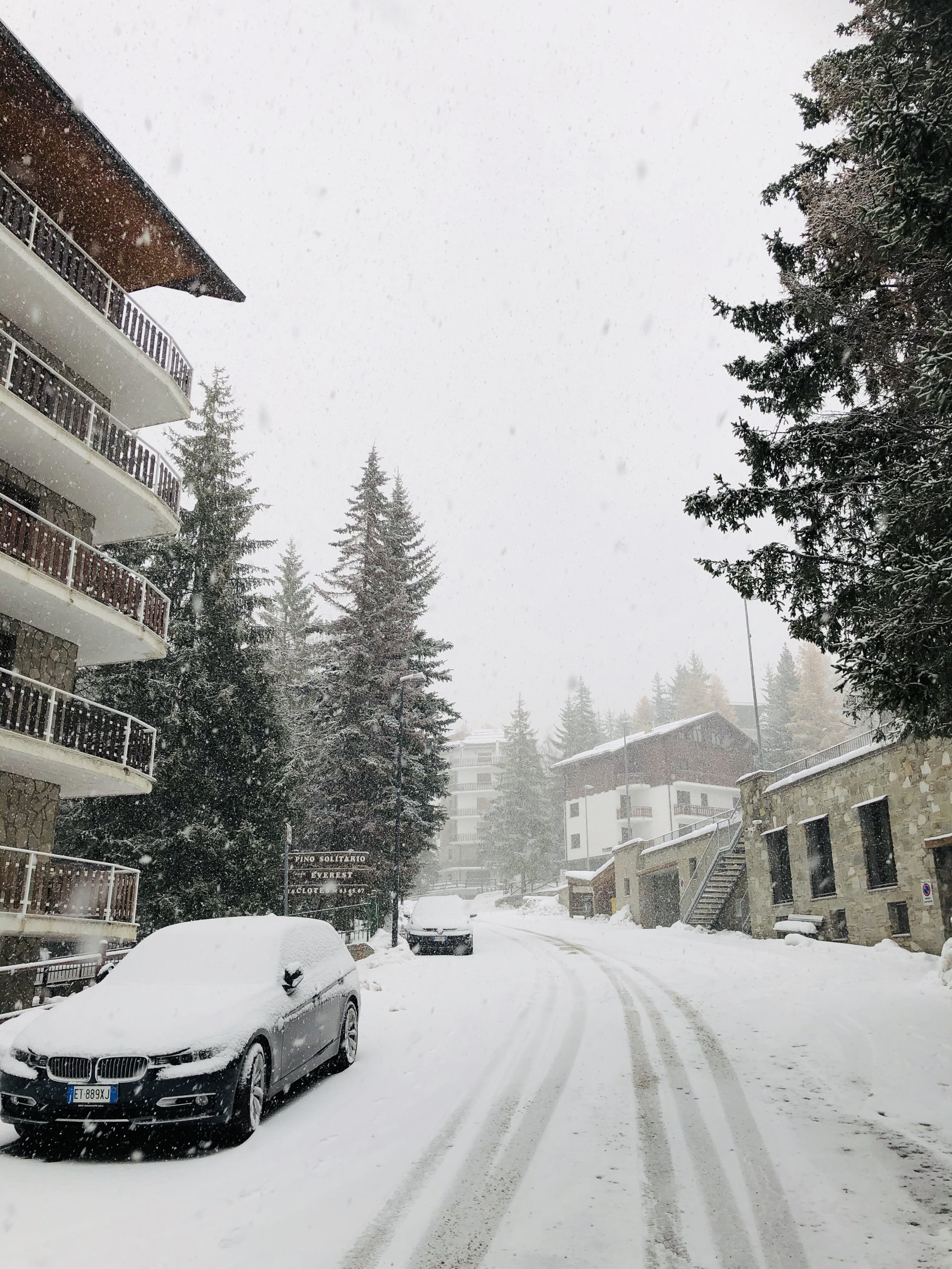 A snow-covered street in an alpine village during a snowfall, with a car parked in the foreground and buildings on either side.