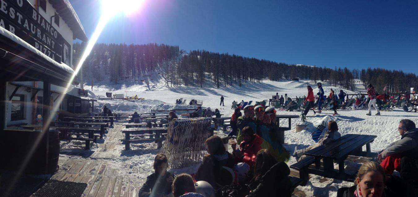 People relaxing on a wooden restaurant terrace on a sunny day at a ski resort, with a snowy mountain slope in the background.