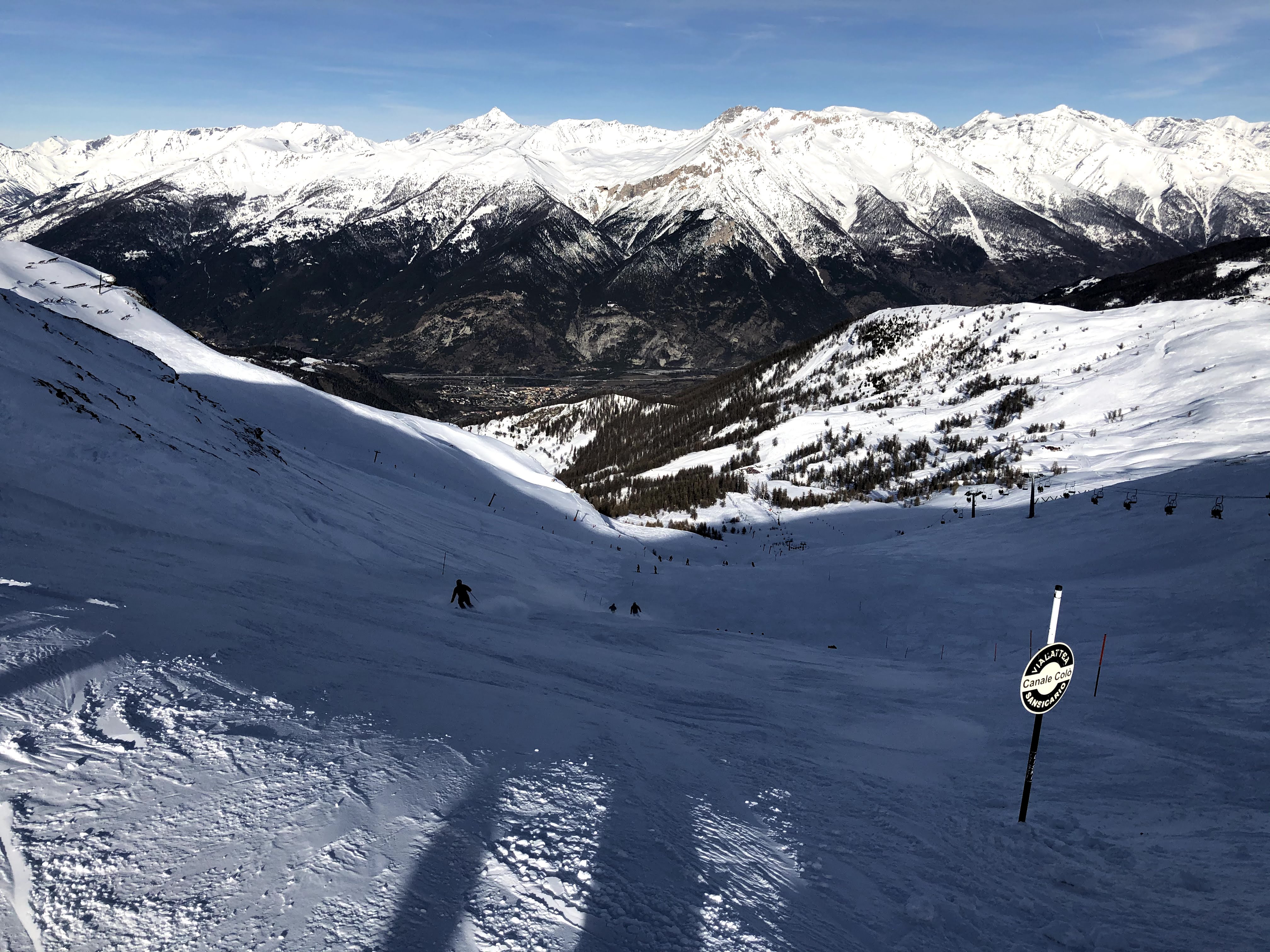 Looking down a steep, snow-covered ski slope towards a valley town with a vast alpine mountain range in the distance.