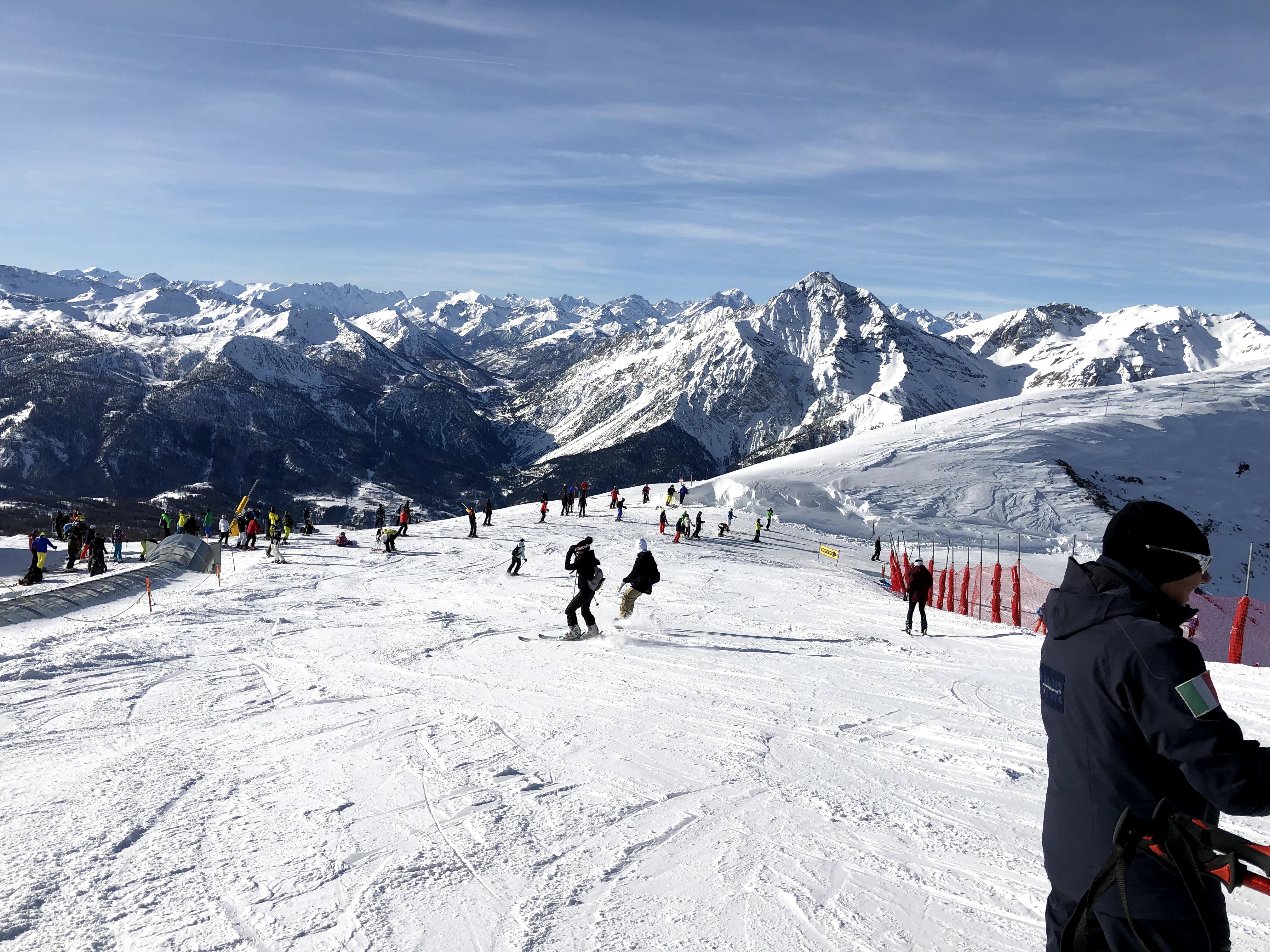 Multiple skiers descending a wide, sunny ski slope with panoramic views of jagged snow-capped mountains in the background.