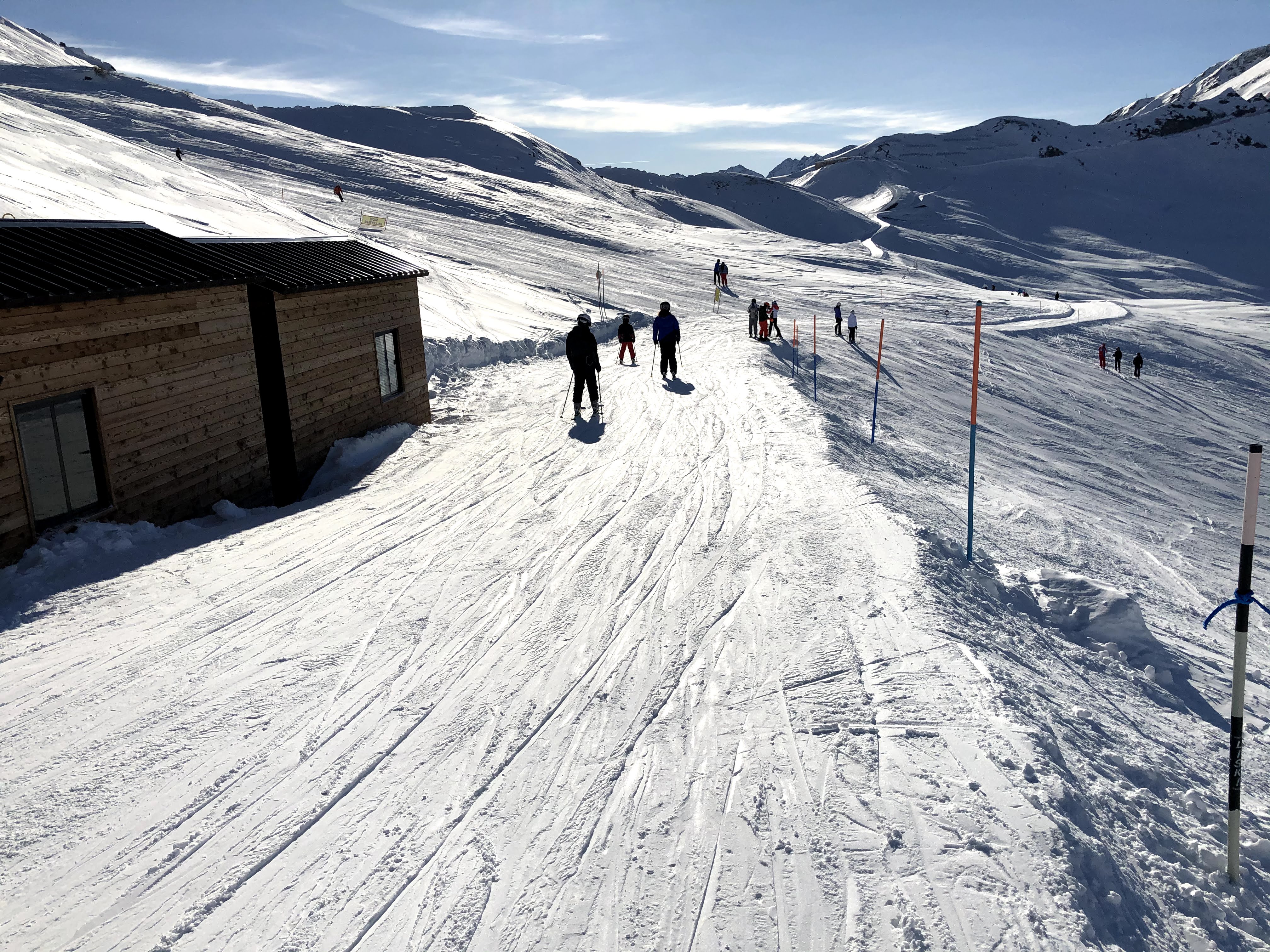 A wooden mountain hut next to a snowy ski trail with several skiers moving along the path under a blue sky.