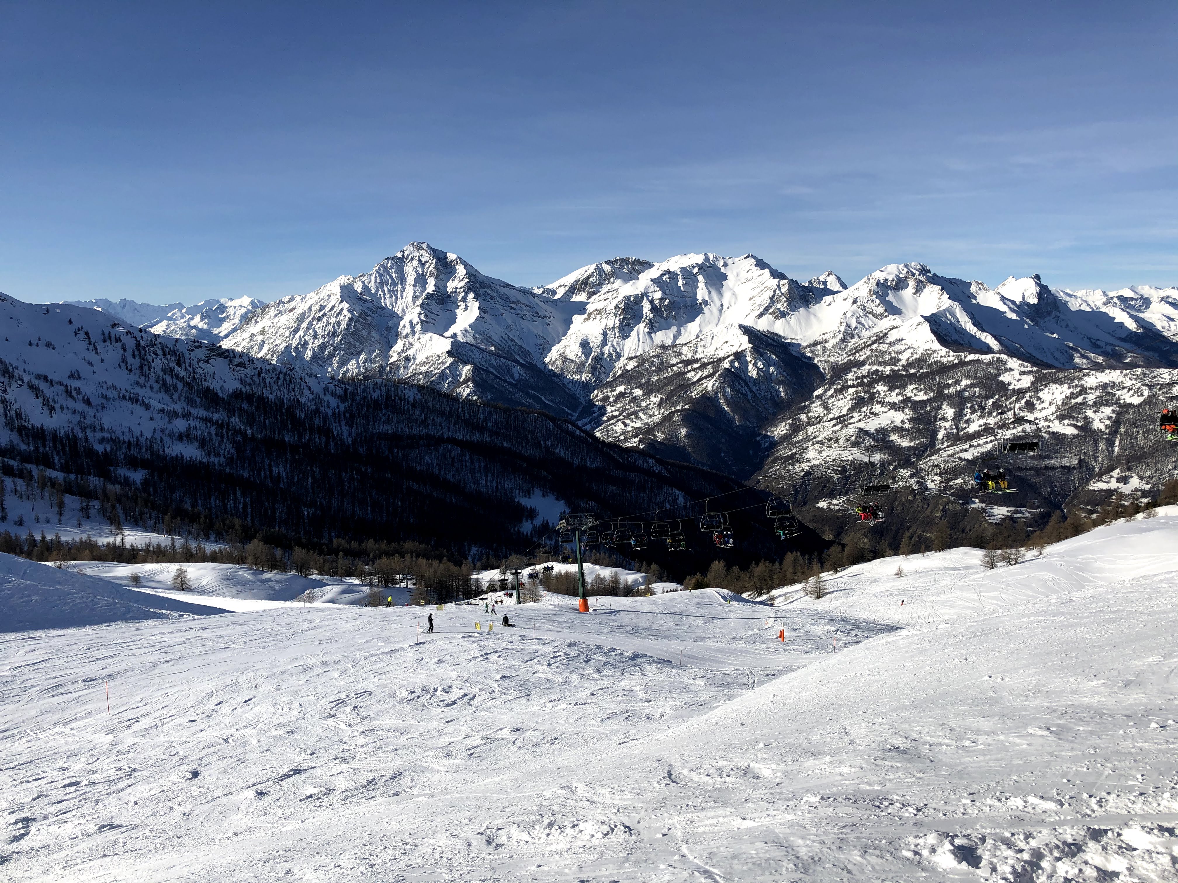 A chairlift operating over a snow-covered ski slope with a sweeping view of snow-capped mountains and pine forests.