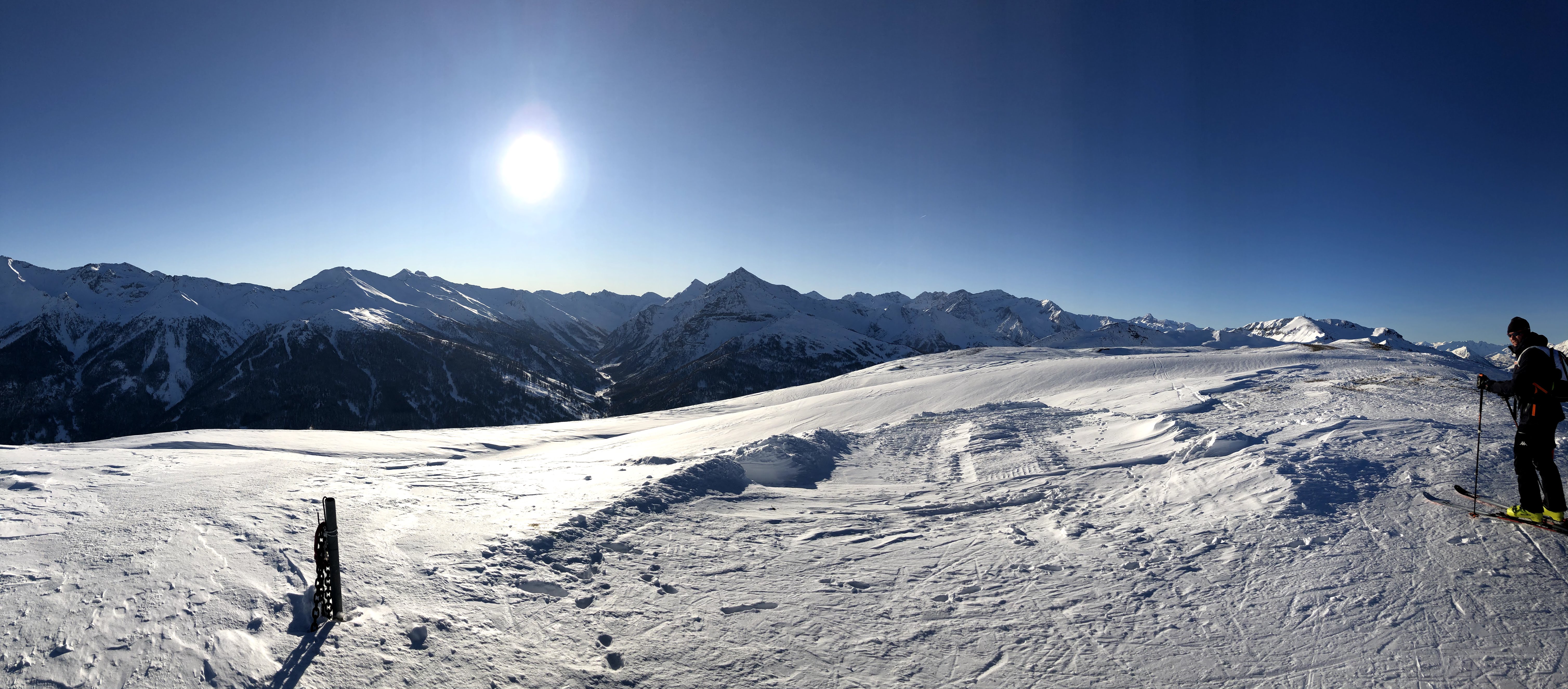 Wide panoramic shot from a snowy mountain ridge with a lone skier on the right, under a bright sun and clear blue sky.