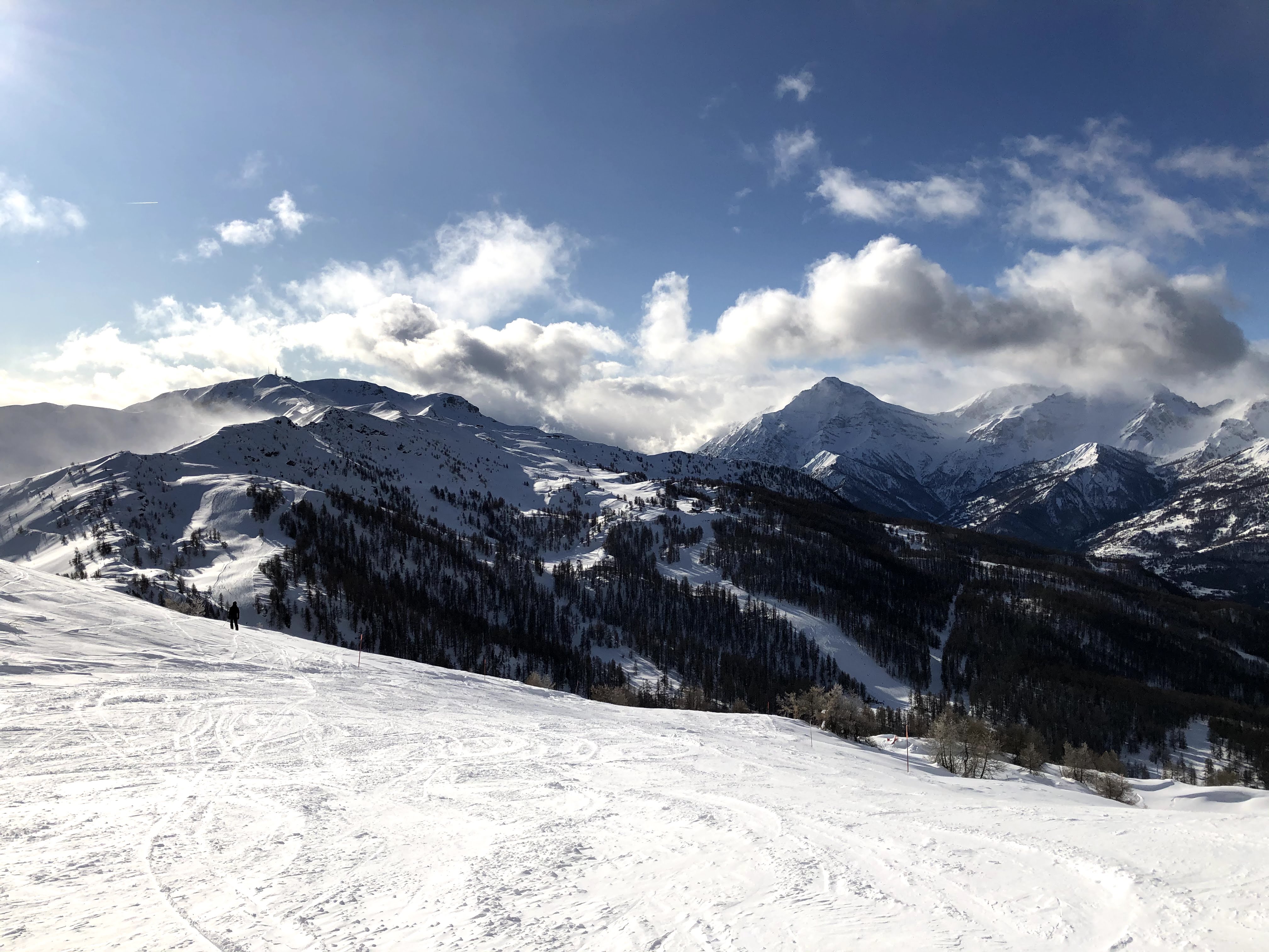 A wide, snow-covered ski slope winding along a pine forest with majestic mountains covered by clouds in the background.