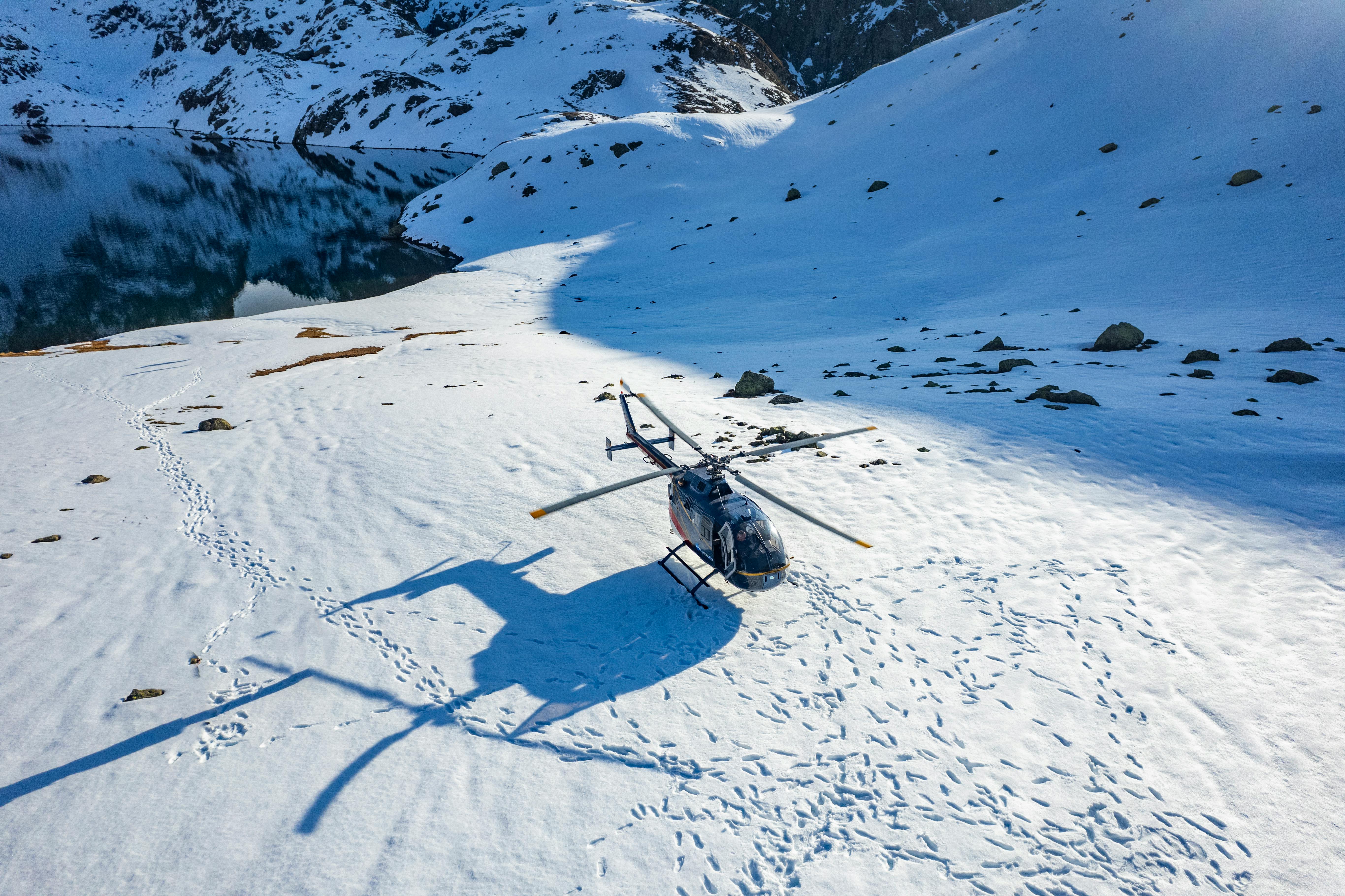 High-angle view of a helicopter parked on a snow-covered alpine landscape next to a dark, partially frozen mountain lake.